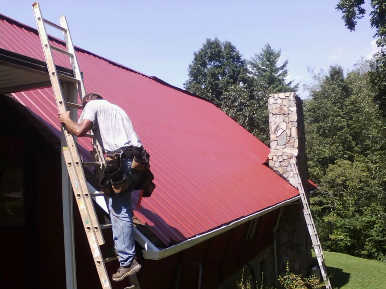 Roofer on a ladder beside a red metal roof and stone chimney — Soffit & Fascia Repair by Roofing Brothers in Olathe